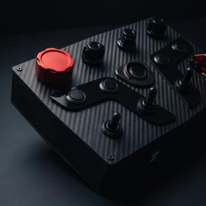 May include: A black control panel with a carbon fiber texture. It features a large red knob, several black toggle switches, and other black control elements. The panel is rectangular and has a dark background.