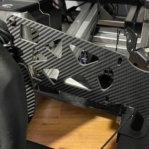 Op de afbeelding: Een carbon fiber sim racing pedaalplaat met uitsparingen en montagehardware. De plaat is gemonteerd op een race simulator rig. Een zwart stuurwiel is zichtbaar op de voorgrond.