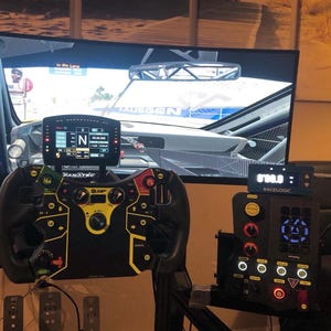 A black racing steering wheel with yellow accents and buttons. The steering wheel is mounted on a stand and connected to a black and gray racing simulator console with a digital display. The console has a carbon fiber pattern and a red button. The screen in the background shows a racing game.