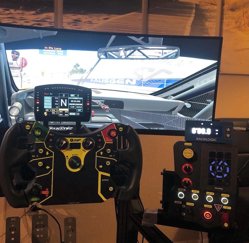 A black racing steering wheel with yellow accents and buttons. The steering wheel is mounted on a stand and connected to a black and gray racing simulator console with a digital display. The console has a carbon fiber pattern and a red button. The screen in the background shows a racing game.