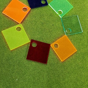 May include: Seven colorful square acrylic tiles with a hole in the center. The tiles are arranged in a circle on a green surface.