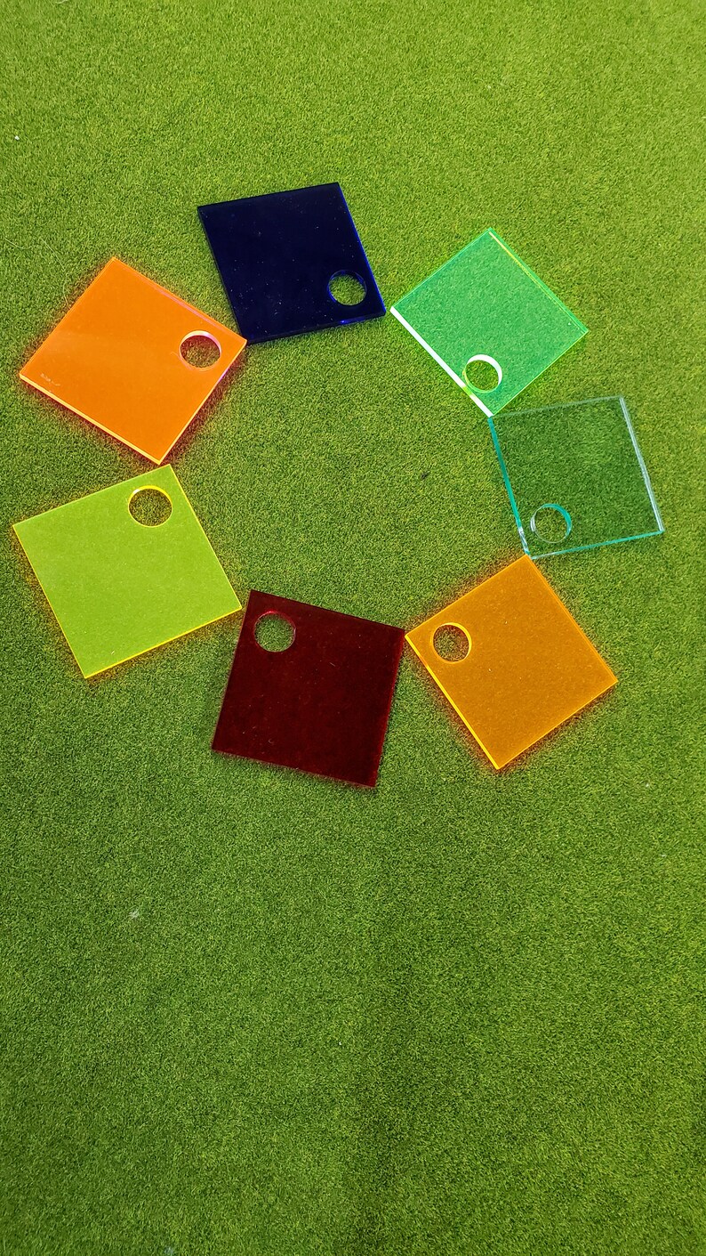 May include: Seven colorful square acrylic tiles with a hole in the center. The tiles are arranged in a circle on a green surface.