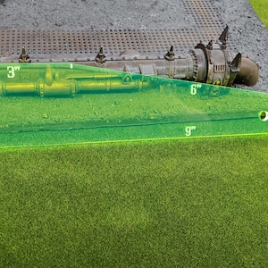 May include: A green ruler with markings in inches, measuring 9 inches long, 6 inches wide, and 3 inches high. The ruler is placed on a brown surface with a metal pipe and other objects.