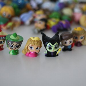 May include: A collection of small plastic figurines of Disney characters, including princesses, villains, and other characters. The figurines are all in a cartoon style and have large eyes. The characters include Cinderella, Maleficent, and Snow White.