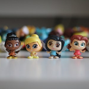 May include: Seven small fairy figurines with wings, each with a different hair color and outfit. The fairies are standing on a white surface.