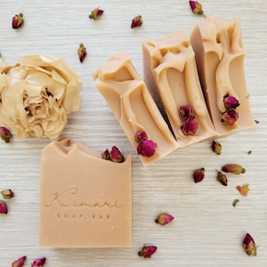 May include: Four bars of light brown soap with a floral scent. The soap bars are arranged in a row, with the top bar slightly overlapping the one below it. The soap bars are decorated with dried rose buds. The soap bars are labelled "Kimari Soap Bar".
