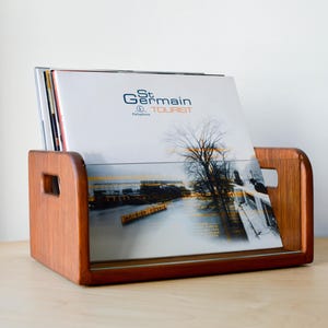 May include: A wooden vinyl record storage crate with cutout handles holding several records. The front-most record features a white cover with the text St Germain Tourist, a graphic of a bare tree, and a river scene in orange and black tones.