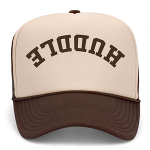 May include: A trucker cap featuring a cream-coloured front panel with the word HUDDLE printed in dark brown block letters in an arched orientation. The cap includes a dark brown brim and a matching dark brown mesh back with a button on top.