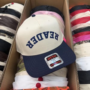 May include: A cream-colored baseball cap with a navy blue brim and a navy blue button on top. The word "READER" is printed in navy blue capital letters across the front panel in an arched design. A rectangular sticker is attached to the brim.
