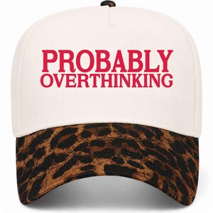 May include: A cream-colored baseball cap featuring the text PROBABLY OVERTHINKING in bold red capital letters on the front panel. The cap has a curved brim with a brown and black leopard print pattern and a small black button on the top crown.