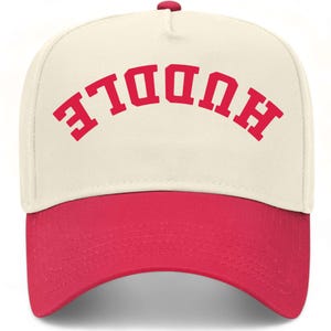 May include: A cream-coloured baseball cap with a red brim and a red button on top. The front panel features the word HUDDLE printed in a bold, red, arched varsity-style font. The cap has a structured five-panel design with a curved visor.