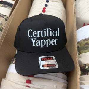 May include: A black baseball cap featuring the white text "Certified Yapper" printed on the front crown. The cap includes a rectangular sticker on the brim with the brand name "OTTO" and the text "Premium Fabric," "Classic Fit," and "Mid Profile."