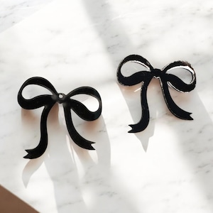 May include: Two black metal bow-shaped ornaments. Each bow has a loop and two flowing tails. The bows are displayed on a white marble surface, with shadows cast across the surface. The bows are likely decorative elements.