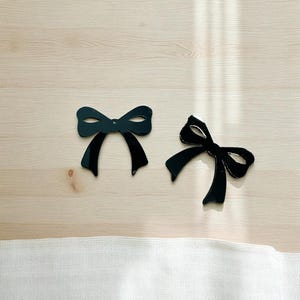 May include: Two black bow-shaped decorations with a glossy finish are displayed on a light wood surface. One bow has a stitched edge. The bows are positioned near a white fabric.