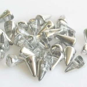 May include: A collection of silver-colored, cone-shaped metal studs with a pointed end and a small hole. The studs are scattered on a white surface, reflecting light and appearing shiny. These are likely used for crafting or embellishment.