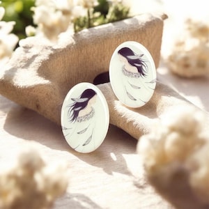 May include: Two oval-shaped earrings with a woman's portrait. The woman has long black hair and is wearing a white dress with feather details. The earrings are white with a glossy finish. The background is a neutral beige.