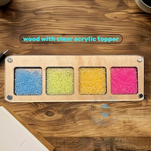 May include: A wooden sensory board with a clear acrylic topper. The board features four square compartments filled with blue, green, yellow, and pink beads. The text "wood with clear acrylic topper" is visible. The logo "NATIVE HANDS CO." is also visible.
