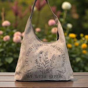 May include: A gray suede shoulder bag with a curved top and a single strap. The bag features an engraved floral design and the text "THEY TRIED TO BURY US. THEY DIDN'T KNOW WE WERE SEEDS."