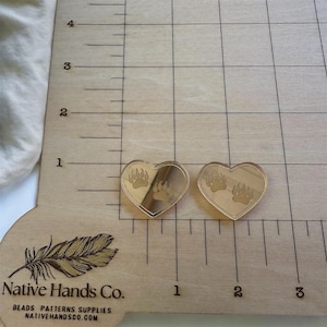 May include: Two heart-shaped, clear acrylic charms with bear paw prints etched on them. The charms are placed on a wooden surface with a grid pattern and the text "Native Hands Co." is visible.