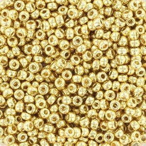 May include: Close-up of a pile of small, round, golden-colored seed beads. The beads are densely packed together, with visible holes in each bead. The surface reflects light, creating a shimmering effect. These beads are likely used for jewelry making or crafts.
