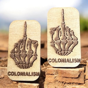 May include: Two wooden plaques with a skeleton hand giving the middle finger. The word "COLONIALISM" is carved at the bottom of each plaque. The plaques are light brown with dark brown detailing.