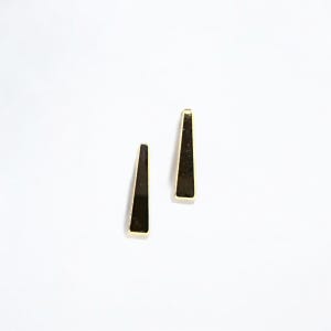May include: Two gold-toned, elongated triangle earrings with a dark, reflective surface. The earrings are set against a plain white background, highlighting their minimalist design and geometric shape. The earrings are a jewelry item.