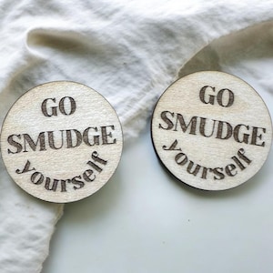 May include: Two round wooden coasters with the phrase "GO SMUDGE Yourself" etched into the surface. The coasters are light brown with dark brown text. One coaster is on a white surface, and the other is on a white cloth.