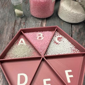 May include: A hexagonal pink plastic bead organizer with six compartments labeled A through F. Each compartment is filled with small, round beads. The beads in compartment A are clear, compartment B is pink, compartment C is white, and the remaining compartments are empty.