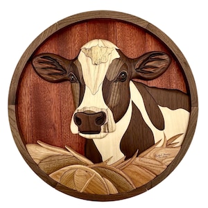 May include: A wooden cow portrait with a brown and white cow head, set against a red-brown background, and framed in a circular wooden border. The cow is resting on a bed of hay, with detailed wood grain patterns.