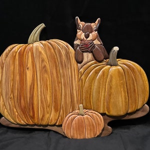 May include: A wooden carving of a squirrel sitting on a pumpkin with two other pumpkins in the background. The pumpkins are carved from wood and have a realistic appearance.