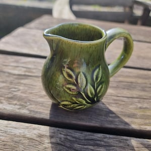 May include: A small, green ceramic creamer with a handle and a spout. The creamer has a textured surface and a leaf design on the front. The creamer is sitting on a wooden surface. This creamer is perfect for serving cream or milk.