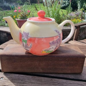May include: A ceramic teapot with a pale yellow upper section and a coral-coloured lower section. The teapot features a matching coral lid and illustrations of two grey tabby cats, one wearing a green bow tie and the other holding a yellow fish.