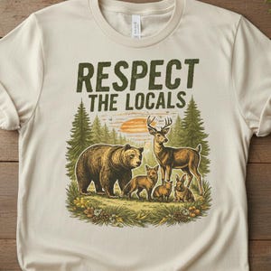 May include: Cream-coloured t-shirt with a forest scene graphic featuring a bear, deer, foxes, and rabbits. The text "RESPECT THE LOCALS" is printed above the illustration in green letters.