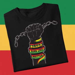 May include: Black t-shirt featuring a clenched fist breaking a chain. The fist is holding a broken chain. The wrist is wrapped with a ribbon that reads "1865 BLACK LIVES MATTER" in red, yellow, and green.