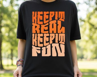 Könnte beinhalten: Schwarzes T-Shirt mit dem Schriftzug "KEEP IT REAL KEEP IT FUN" in orangefarbenen und pfirsichfarbenen, wellenförmigen Buchstaben. Das T-Shirt wird von einer Person gehalten, mit einem verschwommenen grünen Hintergrund.