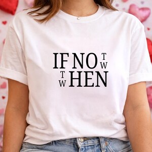 May include: White t-shirt with the text "IF NOT NOW THEN". The text is in a bold, black font. The shirt has short sleeves and a crew neck. The background is a soft pink with heart-shaped decorations.