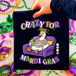 以下が含まれることがあります： 「CRAZY FOR MARDI GRAS」の文字が入ったネイビーブルーのTシャツ。道化師の帽子とマルディグラのビーズを身につけ、紫色のソファでくつろぎ、グリーンのカップを持っている漫画のガチョウが描かれています。