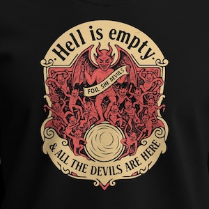 May include: Black t-shirt with a red and beige graphic. The graphic depicts a devil holding a banner reading "FOIL SHE DEVILS" and the text "Hell is empty & all the devils are here."
