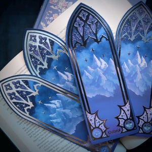 Può includere: Quattro segnalibri blu e argentati con un design ad arco gotico e una scena di montagna ad acquerello. I segnalibri presentano un cielo notturno stellato e il testo "Velacid".
