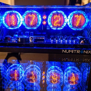 Könnte beinhalten: Nahaufnahme einer Uhr im Vintage-Stil mit vier Nixie-Röhren, die die Zeit 07:27 anzeigen. Die Uhr ist in einem durchsichtigen Acrylgehäuse mit blauer LED-Beleuchtung untergebracht. Die Uhr ist mit "NUMITRONIX" beschriftet.