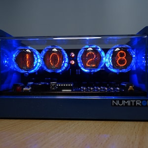 Könnte beinhalten: Eine blaue Uhr mit einem klaren Acrylgehäuse zeigt die Zeit 10:28 mit vier orangefarbenen Nixie-Röhren an. Die Uhr ist mit blauen LEDs beleuchtet und trägt den Markennamen "NUMITRONIX" auf der Vorderseite.