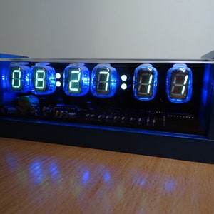 Könnte beinhalten: Eine blaue Uhr mit einem durchsichtigen Acrylgehäuse, das die Zeit 08:27:17 mit grünen digitalen Nixie-Röhren anzeigt. Die Uhr ist mit blauem Licht beleuchtet.