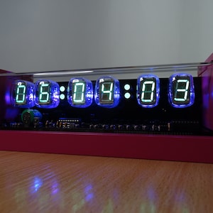 Könnte beinhalten: Eine rosa Uhr mit einem klaren Acrylgehäuse zeigt die Zeit 06:04:08 mit grünen digitalen Ziffern auf schwarzem Hintergrund an. Die Uhr wird mit blauem Licht beleuchtet.