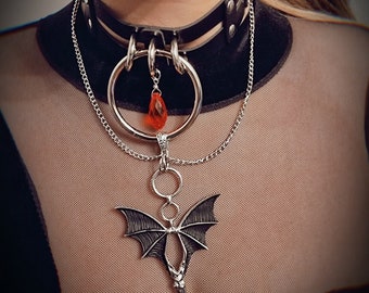 Może przedstawiać: Czarny skórzany choker ze srebrnymi elementami, w tym dużym kółkiem z wiszącym czerwonym kryształem w kształcie łzy. Srebrny łańcuszek łączy się z zawieszką w kształcie nietoperza z rozpostartymi skrzydłami, zawieszoną na kółku.