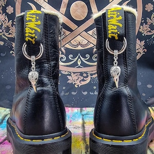 Könnte beinhalten: Schwarze Leder-Plateau-Stiefel mit gelben Nähten und weißem Pelz-Futter. Die Stiefel haben einen silbernen Anhänger an den Schnürsenkeln, auf dem "Dr. Martens" steht.