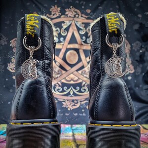 Könnte beinhalten: Ein Paar schwarze Lederstiefel mit gelben Nähten und einem gelben und schwarzen Riemen mit dem Wort "Witch" darauf. Jeder Stiefel hat einen silbernen Anhänger mit einem Eulen- und einem Halbmondmotiv.