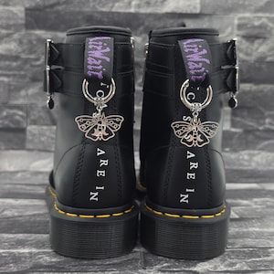 Könnte beinhalten: Ein Paar schwarze Lederstiefel mit gelben Nähten entlang der Sohle, silberfarbenen Schmetterlingsanhängern an den Fersenschlaufen und dem vertikal in Weiß auf die Rückseite jedes Stiefelabsatzes gedruckten Text "ARE IN".