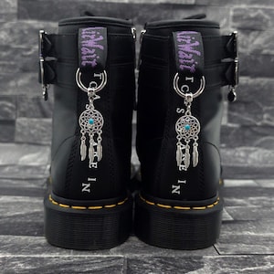 May include: A pair of black leather boots featuring yellow stitching along the sole, silver buckles, and purple heel loops with the text "AirWair". Each boot is adorned with a silver dreamcatcher charm with a turquoise centre bead.