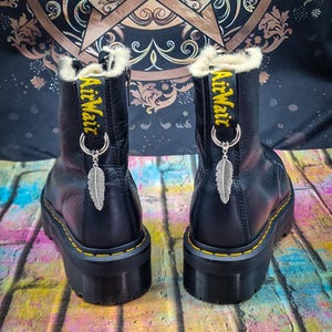 Könnte beinhalten: Schwarze Leder-Plateau-Stiefel mit gelben Nähten und einem weißen Kunstfellfutter. Die Stiefel haben einen silbernen Anhänger mit einer daran befestigten Feder. Der Text "AirWair" ist auf der Rückseite jedes Stiefels gestickt.