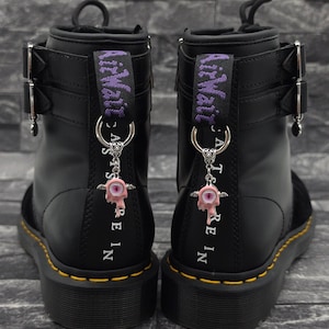 Könnte beinhalten: Ein Paar schwarze Lederstiefel mit zwei silbernen Schnallen an der Seite, gelben Nähten entlang der Sohle und einer violetten Fersenzuglasche mit dem Text AirWair. Ein silberner Ring hält einen rosa, tropfenden Augenanhänger mit Flügeln.